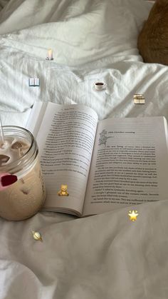 an open book sitting on top of a bed next to a jar of liquid and a cat