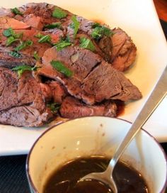 a white plate topped with meat next to a bowl of sauce and a spoon on top of a table