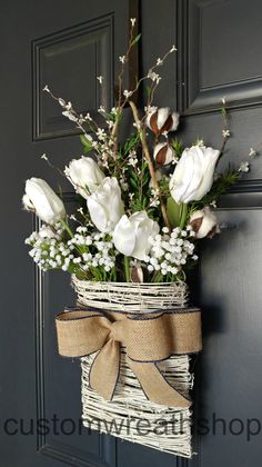 white flowers are in a basket hanging on the front door with burlap ribbon