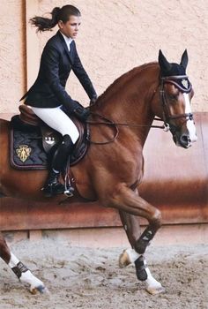 a woman riding on the back of a brown horse next to a building and wearing a black jacket