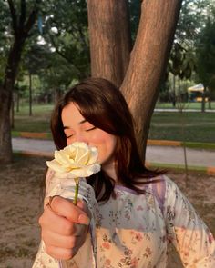 a woman holding a white rose in her right hand while standing next to a tree