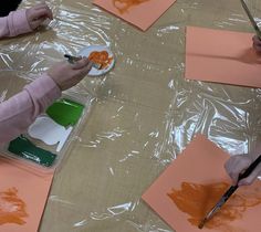 two children are painting with orange paint on paper and plastic wrap around the table, while another child is holding a brush