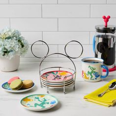 mickey mouse coasters, plates and cups on a white counter with flowers in the background