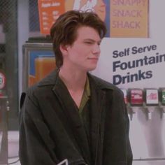 a young man standing in front of a vending machine at a convenience store, wearing a black jacket and green shirt