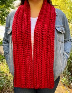 a woman wearing a red knitted scarf in front of some trees and bushes with her hands on her hips