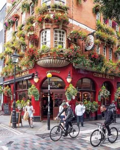 two people riding bikes down the street in front of a building with flowers on it