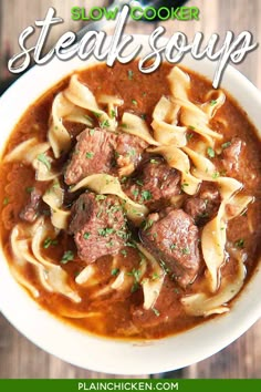 a white bowl filled with pasta and meat soup on top of a wooden table next to a spoon