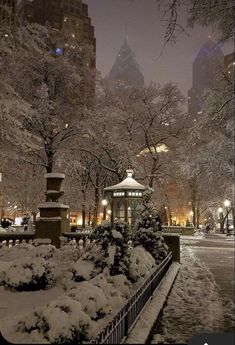 a park covered in snow at night with trees and buildings behind the gazebo on a snowy day