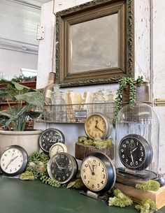 there are many clocks on the table with plants in vases and an old frame