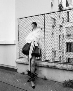 black and white photograph of woman in dress with flowers on the side of street next to fence