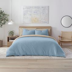 a bed with blue sheets and pillows in a white room next to a chair, mirror and potted plant