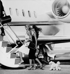 two women walking towards an airplane with a small dog on the ground next to them