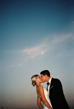 a bride and groom kissing in front of the moon at their beach wedding reception,