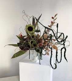 an arrangement of flowers in a vase sitting on a white pedestal against a white wall