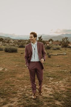 a man in a suit and tie standing on the grass with mountains in the background