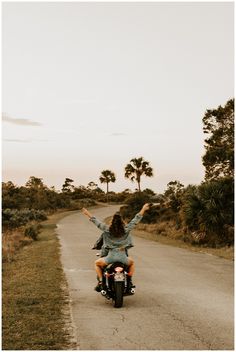 a woman riding on the back of a motorcycle down a country road with palm trees in the background