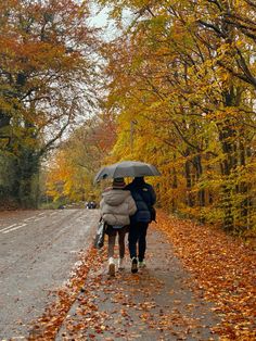 two people walking down the road with an umbrella over their heads in the fall leaves