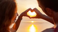 a man and woman making a heart shape with their hands on the beach at sunset