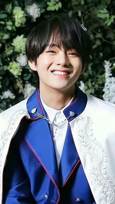 a young man wearing a blue and white jacket smiling at the camera with greenery behind him