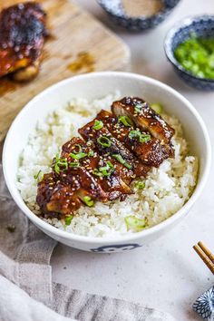 a white bowl filled with rice and meat on top of a wooden cutting board next to chopsticks