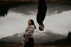 a woman standing next to a man upside down in front of a lake with mountains behind her