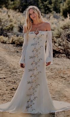 a woman in a long white dress standing on a dirt road with trees and bushes behind her