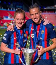 two women in blue and red uniforms holding a silver cup with their arms around each other