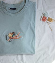 a t - shirt with an embroidered cupid on it next to thread and scissors