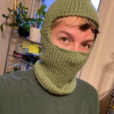 a person wearing a green knitted hat and neck scarf over their face, with a plant in the background