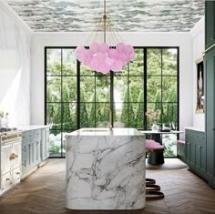 a marble island in the middle of a kitchen with green cabinets and large windows that look out onto trees