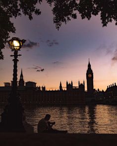 a person sitting on a bench next to the water at dusk with a clock tower in the background