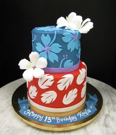 a three tiered birthday cake with white flowers on the top and blue, red, and pink designs