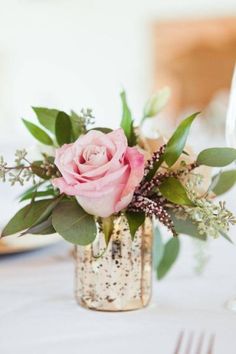 a pink rose in a gold vase on a white table cloth with greenery and silverware