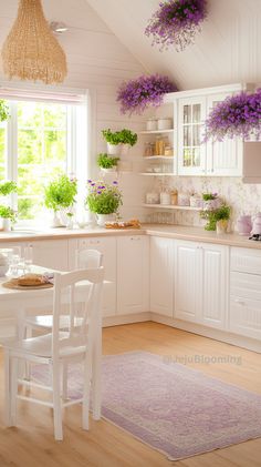 a kitchen filled with lots of white furniture and purple flowers hanging from the ceiling above