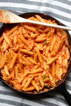 a skillet filled with pasta on top of a striped table cloth next to a wooden spoon