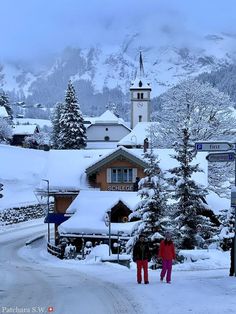 two people are walking in the snow near some buildings and trees with mountains in the background