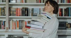 a woman holding a stack of books in front of a book shelf filled with books