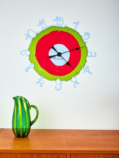 a green and red clock sitting on top of a wooden table next to a vase