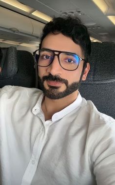 a man with glasses and a beard is sitting in an airplane looking at the camera