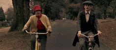 two people riding bikes down a street with trees in the backgrouns and one person wearing a red hat