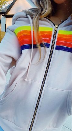 a woman with blonde hair wearing a rainbow colored hoodie and blue jeans standing in front of a house