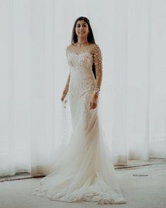 a woman standing in front of a window wearing a wedding dress with long sleeves and beading