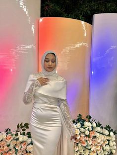 a woman standing in front of flowers wearing a white dress and head scarf with long sleeves