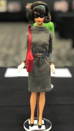 a doll is wearing a dress and holding a red purse on a black surface with other dolls in the background