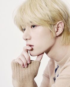 a young man with blonde hair wearing a beige sweater and holding his hand on his chin