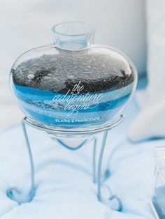 a glass bottle with some black sand in it sitting on a metal stand next to a pillow