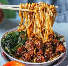 someone holding chopsticks over a bowl of noodles with meat and spinach in it