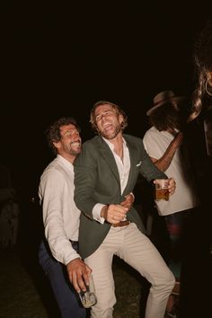 two men are laughing and having fun at an outdoor party in the dark, one is holding a beer