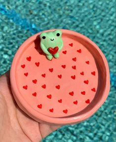 a small frog sitting on top of a pink bowl next to a pool with hearts