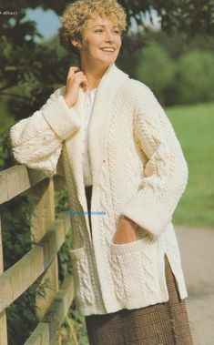 a woman standing next to a wooden fence wearing a white cardigan sweater and brown skirt